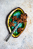 Star anise from above on a ceramic bowl in bright shades of green and yellow with a spoon resting on a textured grey surface.
