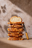 A stack of artisan bread slices with a crispy texture lies elegantly on a rustic surface A knife with a wooden handle leans casually against the stack, creating a cosy, homely atmosphere