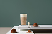 A glass of coffee with milk cream, surrounded by coffee capsules Brown sugar falls from a spoon nearby, adding texture to the minimalist scene with the blue-green background