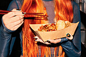 A woman with long orange hair holds a paper tray of Asian street food and eats with chopsticks. She is in a bustling food market enjoying delicious oriental flavours