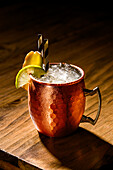 A refreshing Moscow Mule, served in a classic copper mug, garnished with fresh lime and ginger The iced drink sparkles under the warm lighting, emphasising its inviting texture and vibrant appearance
