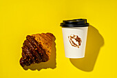 Top view of a decadent chocolate croissant with a takeaway coffee cup showing a lipstick stain, all against a bright yellow background.