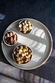 Top view of a round, textured tray with three bowls containing different nuts. Each tray contains a different type of nut: almonds, hazelnuts and peanuts, neatly arranged on a dark surface that highlights the textures and natural colours of the nuts.