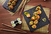 Top view of a range of sushi, including cucumber maki and spicy tuna rolls, presented on a black slate, accompanied by ginger, wasabi, chopsticks and bamboo mats on a wooden table.