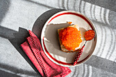 Top view of a vibrant dish of healthy organic red trout caviar, served on sliced bread, on a white and red plate, accompanied by a napkin and a spoon, all under natural sunlight.