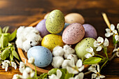 A colourful selection of pastel Easter eggs lies between delicate white flowers and soft cotton on a wooden surface, creating a festive and elegant spring atmosphere.