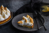 A slice of homemade lemon cake lies elegantly on a dark-coloured plate, accompanied by a fork and knife. A glass with a slice of lemon adds a refreshing touch to the minimalist scene.