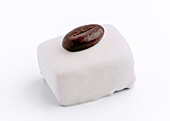 A delicate, square petit four is decorated with a single coffee bean on a white glaze. This gourmet dessert combines sophistication and simplicity in one bite.