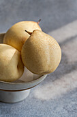 Three ripe white Chinese pears rest in a clean ceramic bowl. The soft shade and textured background emphasise the freshness and simplicity of the scene. Perfect for culinary themes.