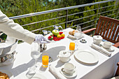 Elegant waitress with white gloves sets a luxurious outdoor breakfast table with fresh fruit, pastries and drinks. Scenic view with greenery in the background