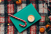 Top view of a festive Christmas decoration with a lit candle, a candy cane, a book and fresh mandarins on a chequered background, capturing the warmth and spirit of the Christmas season.