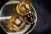 Top view of two spiced cognac-based cocktails with cinnamon and star anise, served alongside a bowl of mixed nuts, including almonds and peanuts, on a dark textured surface.