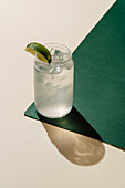 A chilled glass of classic lemonade, served with ice cubes and a slice of lime. The drink stands on a two-tone, green and cream-coloured surface and casts a soft shadow.