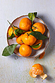 Ein lebhaftes Arrangement frischer Mandarinen mit grünen Blättern, die auf einem rustikalen Teller vor einem strukturierten Hintergrund präsentiert werden. Eine Mandarine ist geschält, so dass die saftigen Segmente zum Vorschein kommen.