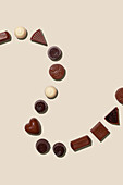 Top view of a collection of different chocolate truffles, skilfully arranged on a soft beige background. Contains heart-shaped, round and geometric pieces with a visible creamy texture and cocoa dust.