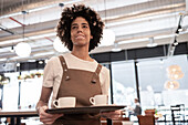 A friendly café employee with vitiligo in a brown apron carries a tray with two coffee cups, demonstrating the excellent service in a bright, modern café with large windows and stylish lighting