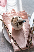 Viewed from above, an artfully arranged Easter table shows a porcelain bowl with speckled eggs adorned with delicate feathers next to a pastel pink napkin and elegant silver cutlery in the soft daylight.