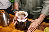 A barista skilfully prepares coffee specialities using the pour-over method. The equipment consists of a stainless steel boiler and a glass carafe on a digital scale that emphasises precision.
