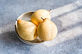 Three white Chinese pears in a bowl are placed on a textured surface. The smooth, speckled skin of the pears contrasts with the rough background and captures natural light and shadow.