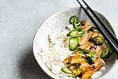 Top view of a hearty dish of grilled tofu slices and white rice, garnished with fiery green chilli slices, served in a speckled bowl with black chopsticks.