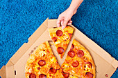 A hand reaches for a slice of pepperoni pizza from a box on a fluffy blue rug that creates a relaxed and cosy atmosphere, perfect for an evening at home