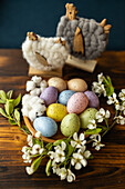 A rustic ambience with colourful speckled Easter eggs in a wooden bowl, surrounded by white flowers and woolly chickens on a wooden table.