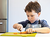 A little boy in an apron with a star pattern concentrates on cutting a carrot with a small knife on a green chopping board in a modern kitchen.