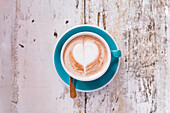 A cappuccino in a turquoise-coloured cup with a heart-shaped latte art design. The cup stands on a rustic white wooden table and creates a cosy and artistic atmosphere.
