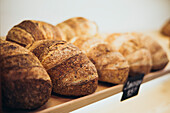 A selection of freshly baked loaves of bread arranged on wooden shelves in a bakery. The rustic crusts show the artisan quality and warmth of traditional baking