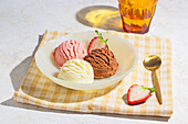 A close-up of three scoops of vanilla, chocolate and strawberry ice cream in a bowl, shot in soft natural light with a high-key aesthetic