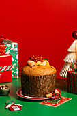 A beautifully decorated panettone lies on a bright green surface with a festive red background. It is surrounded by wrapped presents, Christmas tree decorations and cinnamon sticks