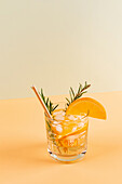 An inviting cocktail garnished with orange slices and sprigs of rosemary, served on ice in a clear glass against a two-tone pastel-coloured background. Perfect for a summery refreshment.