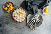 Top view of a homemade lemon cake with golden peaks, sliced lemons, antique cutlery and a dark fabric. The scene is reminiscent of a rustic, cosy kitchen.