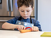 A focussed child in an apron with a star pattern peels a carrot in a bright kitchen environment. The picture shows concentration and learning in a domestic environment.