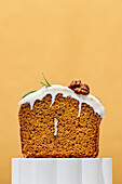 A delicious carrot cake with creamy white icing is set against a warm orange-coloured background. Sprinkled with walnuts and rosemary, this dessert is a perfect treat.