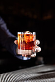 Close-up of an old-fashioned whiskey cocktail held by a hand over a textured bar counter A slice of orange garnishes the drink, contrasting beautifully with its rich amber colour