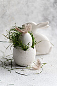 A softly textured Easter egg nestled in bright green grass, adorned with a delicate feather and placed next to a white ceramic bunny. This elegant arrangement captures the joy and renewal that Easter symbolises.