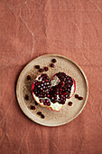 A rustic ceramic plate shows a sliced pomegranate with glittering seeds on a textured, earthy fabric background. The deep red fruit adds vibrancy to the warm surroundings.