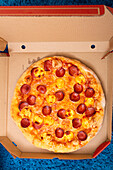 A delicious pizza with salami and cheese lies invitingly in an open box on a blue plush carpet and radiates a cosy, relaxed and homely atmosphere