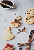 Draufsicht auf selbstgebackene Osterhasenkekse, die mit Hibiskustee kreativ bemalt wurden, gepaart mit einem Pinsel und einigen verstreuten Kekskrümeln auf einer weißen Fläche. Ein festliches und charmantes Backprojekt für den Frühling.