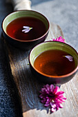 Two ceramic cups filled with tea stand on a textured wooden board Each cup has floating pink petals accented by nearby pink flowers, creating a serene, rustic scene