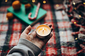 Cropped, unrecognisable hand of a person holding a lit candle against a blurred, festive background with hints of oranges and candy canes, embodying the warmth and cheerfulness of the Christmas season.