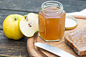 Ein Glas hausgemachter Quittenmarmelade steht auf einem Holzbrett neben frischen Quitten und einer Scheibe Brot. Ein Messer liegt daneben, bereit zum Verteilen. Rustikal und einladend.