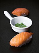 Close-up of fresh salmon sushi and a small bowl of wasabi on a smooth, black stone surface, allowing for a minimalist and elegant presentation of the food.