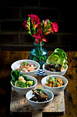 A selection of bowls with Asian dishes arranged on a wooden board The dishes contain vegetables, meat and side dishes A vase of bright red flowers provides a decorative accent in the background