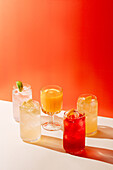 Five different lemonades stand on a surface against a bright orange background. Each glass has different citrus accents and offers a refreshing visual appeal.