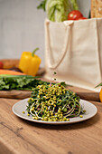 A fresh topping with green sprouts on a white plate stands on a wooden table. In the background, various vegetables and a reusable bag add a lively touch.