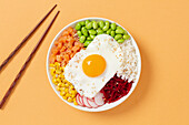 A colourful poke bowl with fresh ingredients such as edamame, corn, salmon, beetroot and a fried egg, elegantly presented with chopsticks against a warm background.