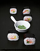 A stylish sushi arrangement on a black stone plate with several pieces of sushi and a white bowl filled with green wasabi. The composition is minimalist and elegant.