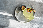 Top view of an elegant cocktail of gin, tonic and passion fruit juice, garnished with a halved passion fruit, served on a metal tray under natural shadow light that captures the essence of a refreshing drink.